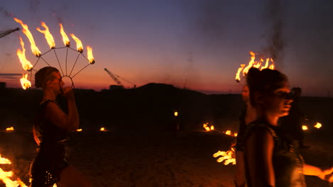 Eine-Gruppe-Von-Männern-Und-Frauen-Zeigt-Nachts-Eine-Feuershow-Auf-Dem-Sand-Vor-Dem-Hintergrund-Von-Feuer-Und-Turmdrehkränen.